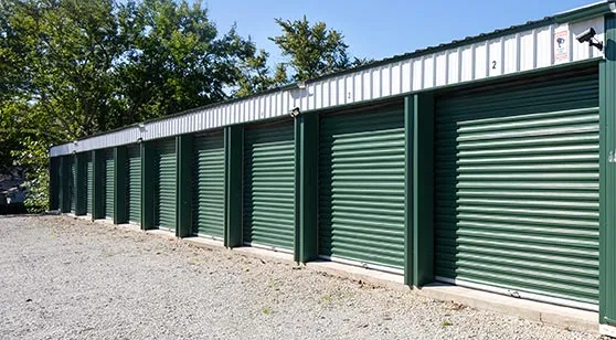 A row of green roll-up doors on a drive-up storage building, with a gravel drive in front and trees behind.
